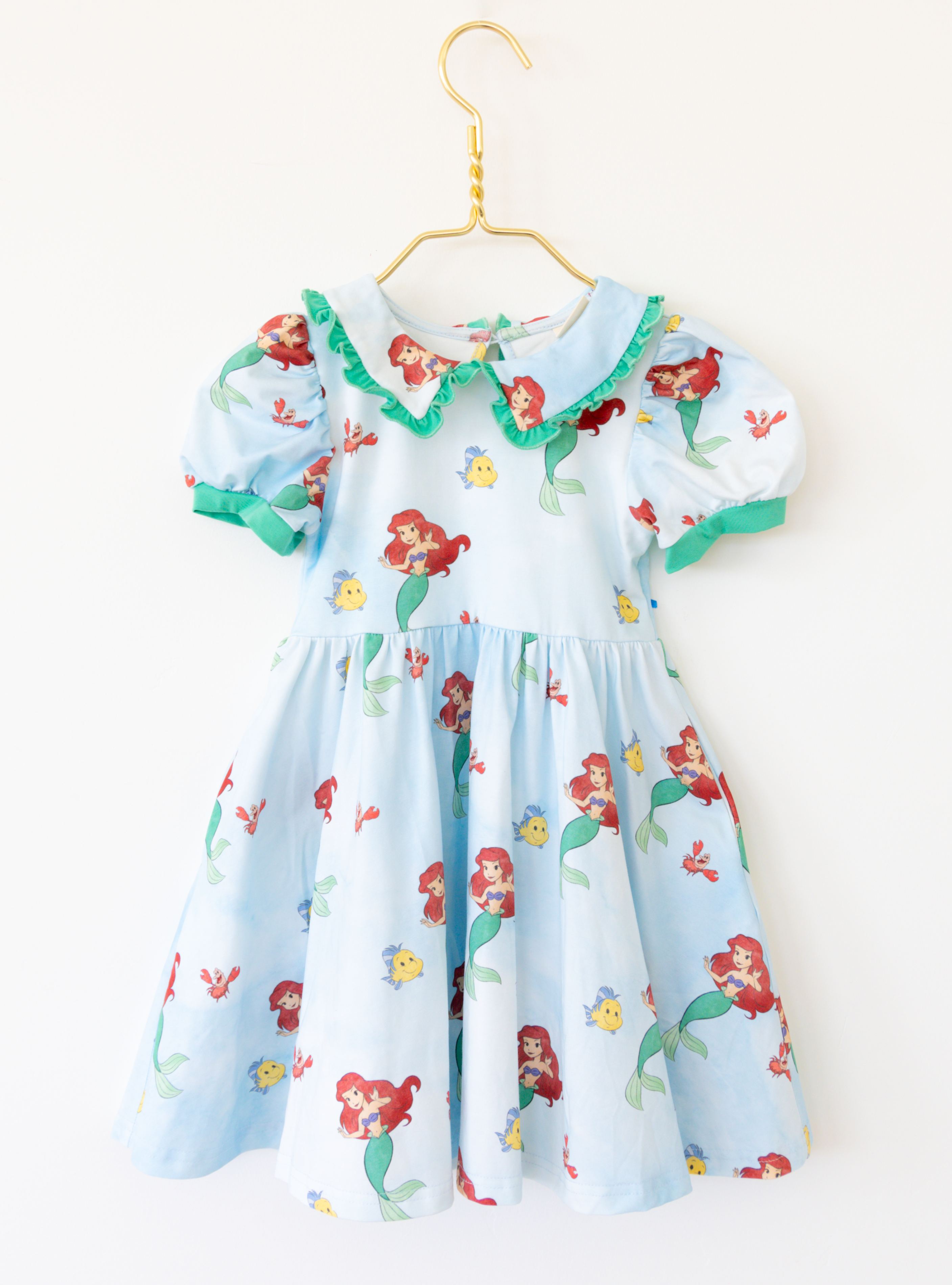 Disney and Taylor Joelle branded Ariel dress with an Ariel, Flounder, and Sebastian print all around. This dress has puff sleeves, and green detailing around the sleeves and collar. 