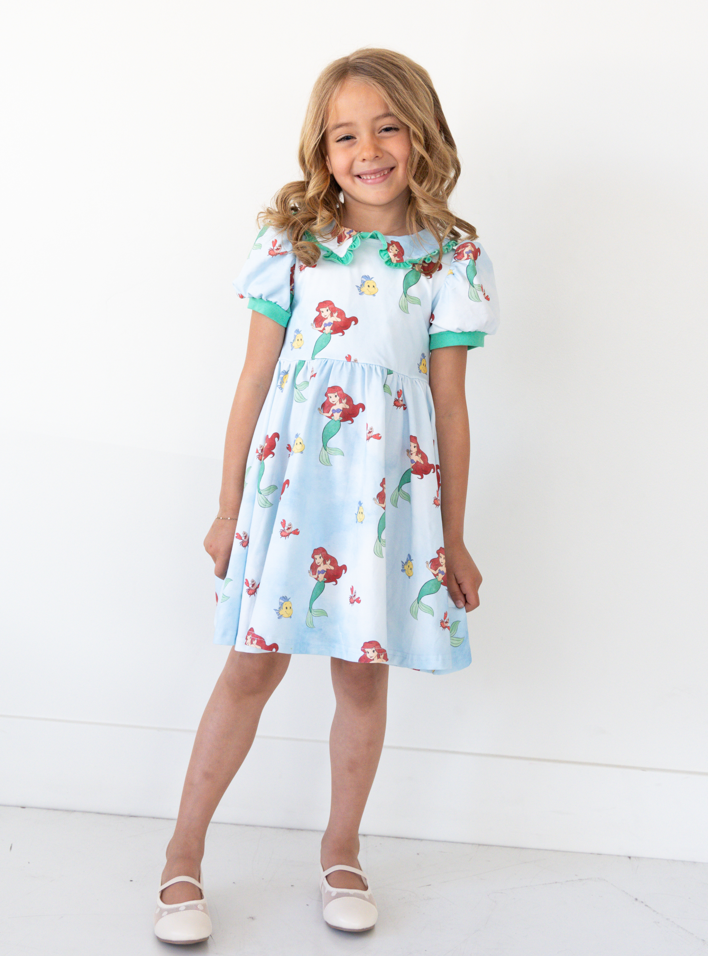 Disney and Taylor Joelle branded Ariel dress with an Ariel, Flounder, and Sebastian print all around. This dress has puff sleeves, and green detailing around the sleeves and collar. 