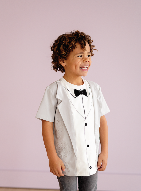 Shirt: White tuxedo-style shirt for boys.  Color: White. Pattern: Tuxedo design. Material: Cotton. Occasion: Formal. Style: Classic. Brand: Taylor Joelle. Image: A young boy wearing a white tuxedo-style shirt. The shirt has a tuxedo design on it, with a black bow tie and buttons. The shirt is made of cotton and has a crew neck. The shirt is short-sleeved. The shirt can be purchased on TaylorJoelle.com